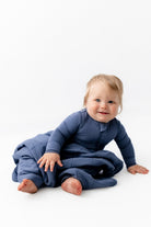 A baby wearing a navy blue long-sleeve footed sleep sack sits on a white studio floor, looking at the camera.