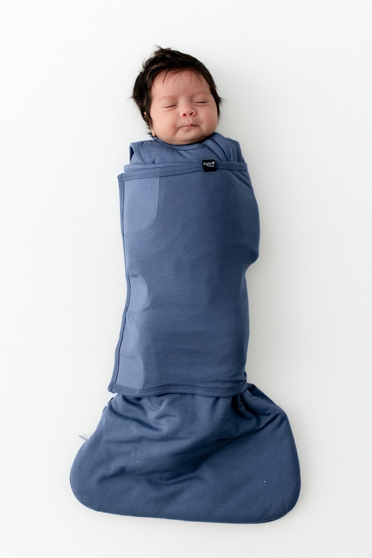 A sleeping newborn baby swaddled in a dusty blue wrap blanket, lying flat on a clean white background. The blanket is secured with a small black brand tag that reads 'MAMmoth'.