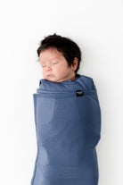 A sleeping newborn baby wrapped in a dusty blue fabric baby carrier wrap, shown against a plain white background.