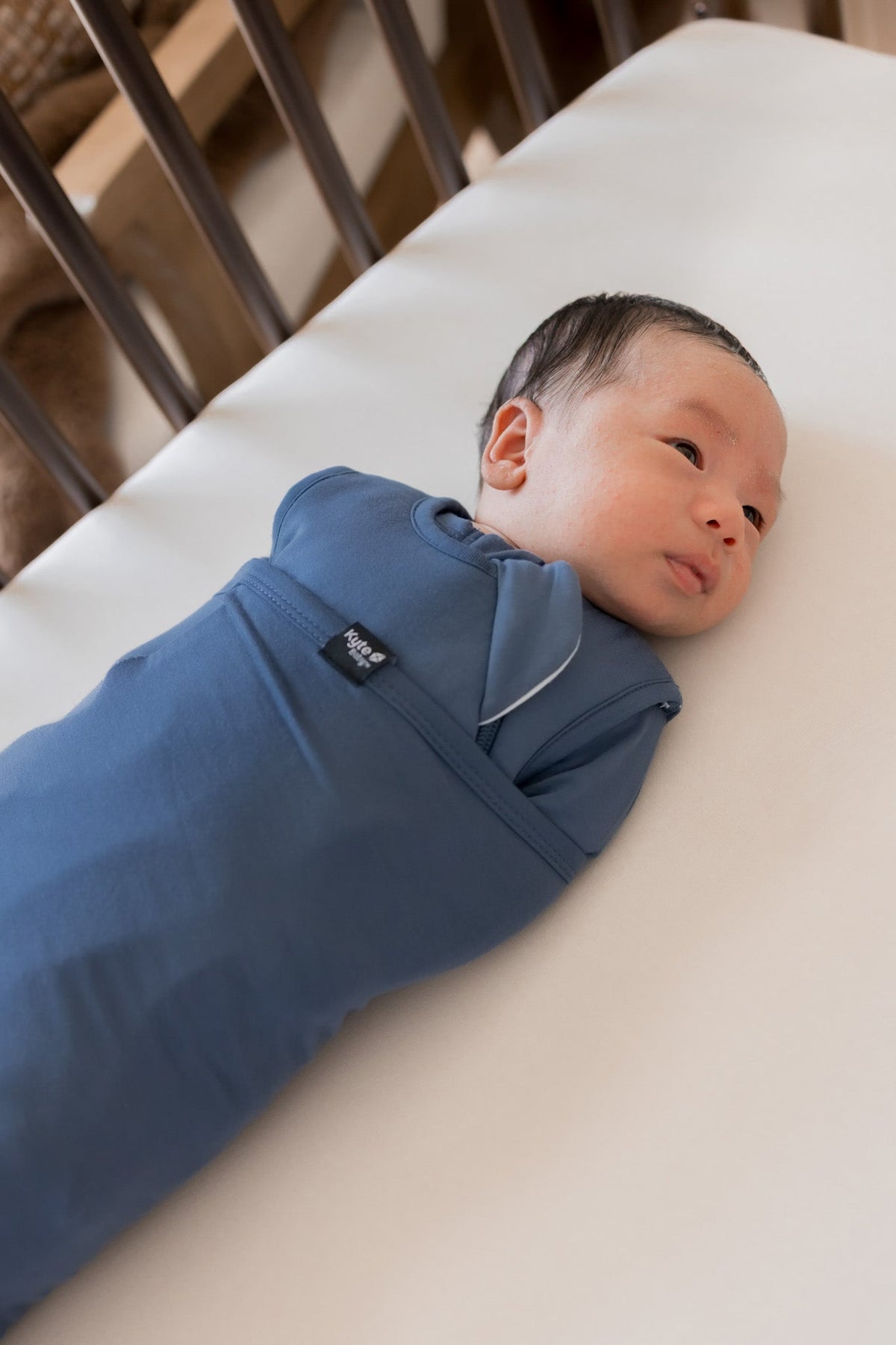 A newborn baby lying on a white mattress, swaddled in a blue wrap blanket. The baby is wearing a matching blue one-piece outfit with a small black brand tag that reads 'baba&boo'.