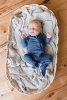 A sleeping newborn baby wearing a navy blue footed one-piece outfit, lying on a textured cream knit blanket inside a woven oval bassinet on a wooden floor.
