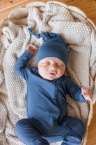A sleeping newborn baby wearing a navy blue one-piece jumpsuit and matching beanie hat, lying on a textured cream knit blanket inside a woven basket.