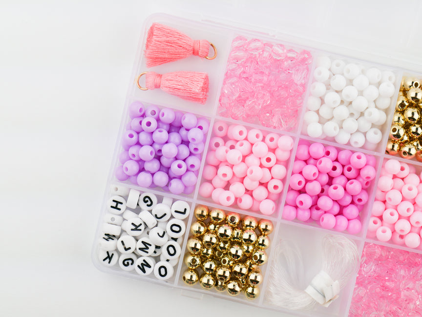 Princess Party DIY Bracelet Kit storage box shown from above, filled with sorted compartments of pink tassels, white and pink round beads, gold spacer beads, alphabet letter beads, and elastic cording.