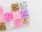 Princess Party DIY Bracelet Kit storage box shown from above, filled with sorted compartments of pink tassels, white and pink round beads, gold spacer beads, alphabet letter beads, and elastic cording.