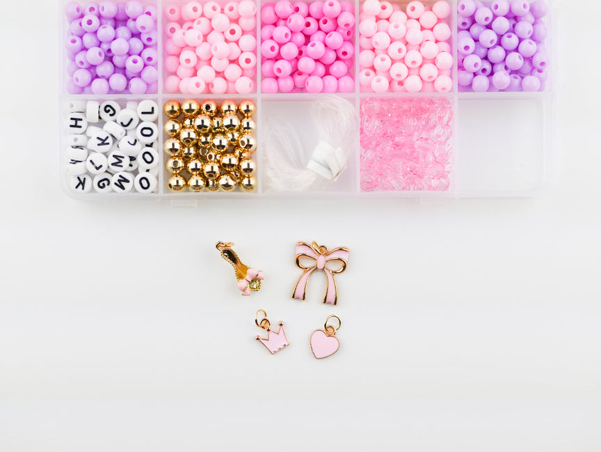 Princess Party DIY Bracelet Kit featuring an open compartment storage box filled with colorful pink, purple, and white round beads, alphabet letter beads, and gold spacer beads, alongside several gold charms including a bow, crown, and heart.