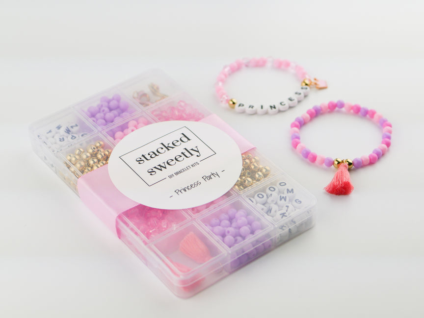 Princess Party DIY Bracelet Kit featuring a compartment storage box with a "stacked sweetly - Princess Party" label, filled with colorful beads, alphabet letter beads, and charms next to two completed pink, purple, and gold beaded bracelets, including one spelling "PRINCESS" with a small crown and tassel charm.