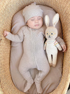 Baby in a textured outfit lying next to a knitted rabbit toy in a woven basket.