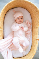 Baby in a pink outfit and hat lying in a wicker crib with white bedding.