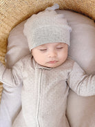 Baby in a beige outfit and hat lying on a cushion