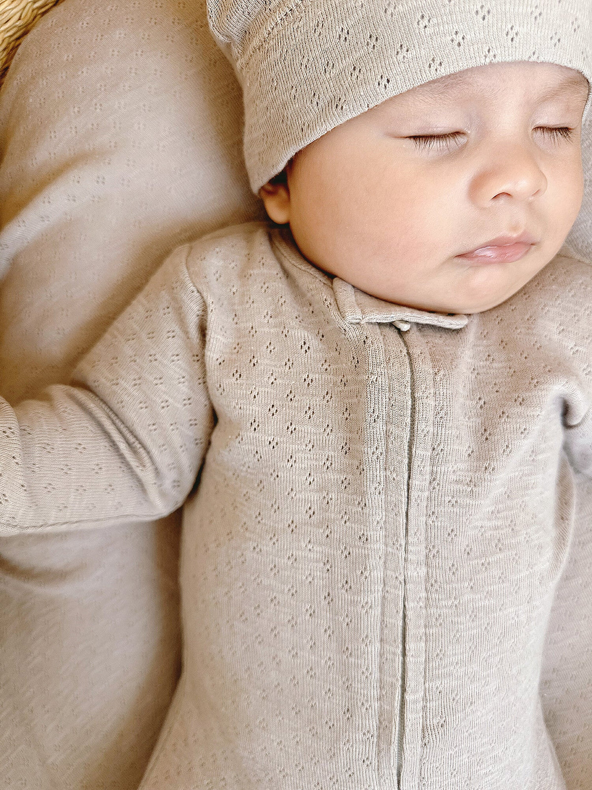 Baby wearing a beige knitted outfit and hat, lying on a soft surface.
