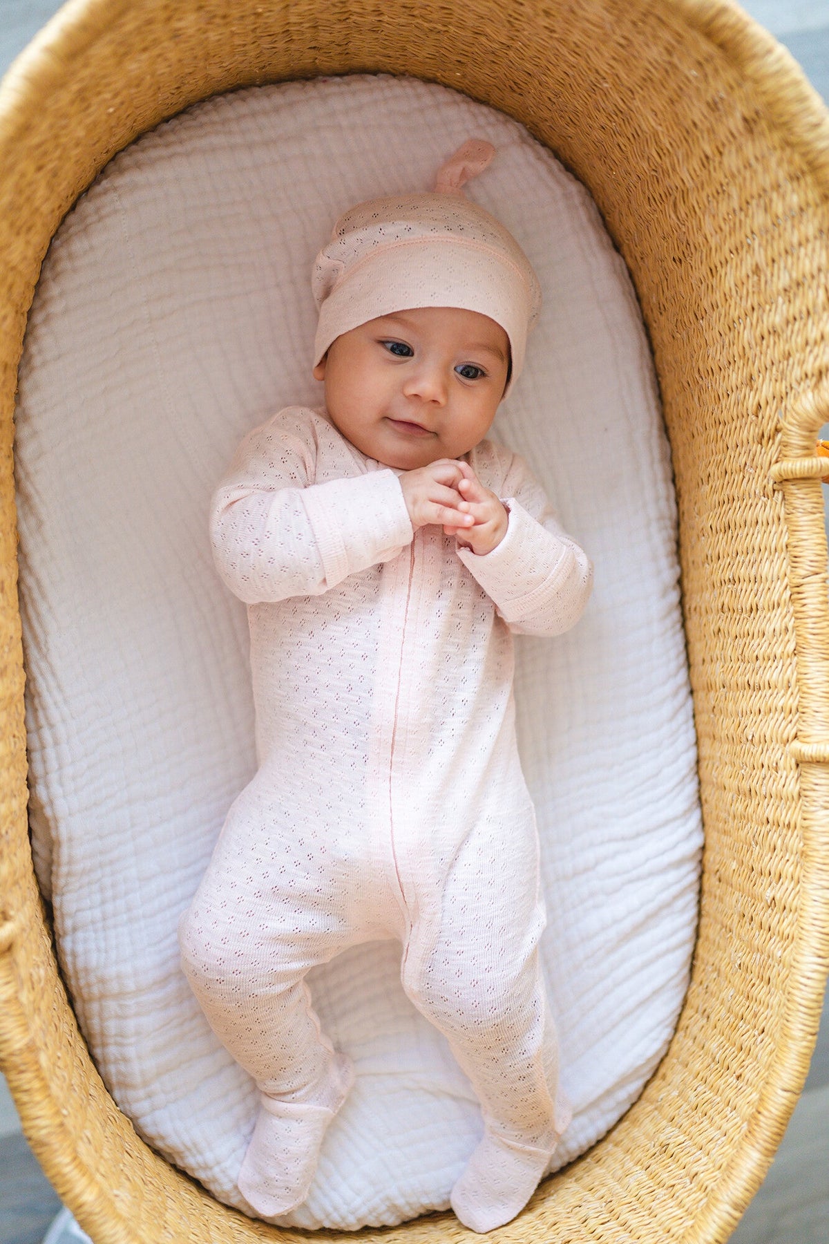 Baby in a pink outfit and hat lying in a woven basket with white bedding.