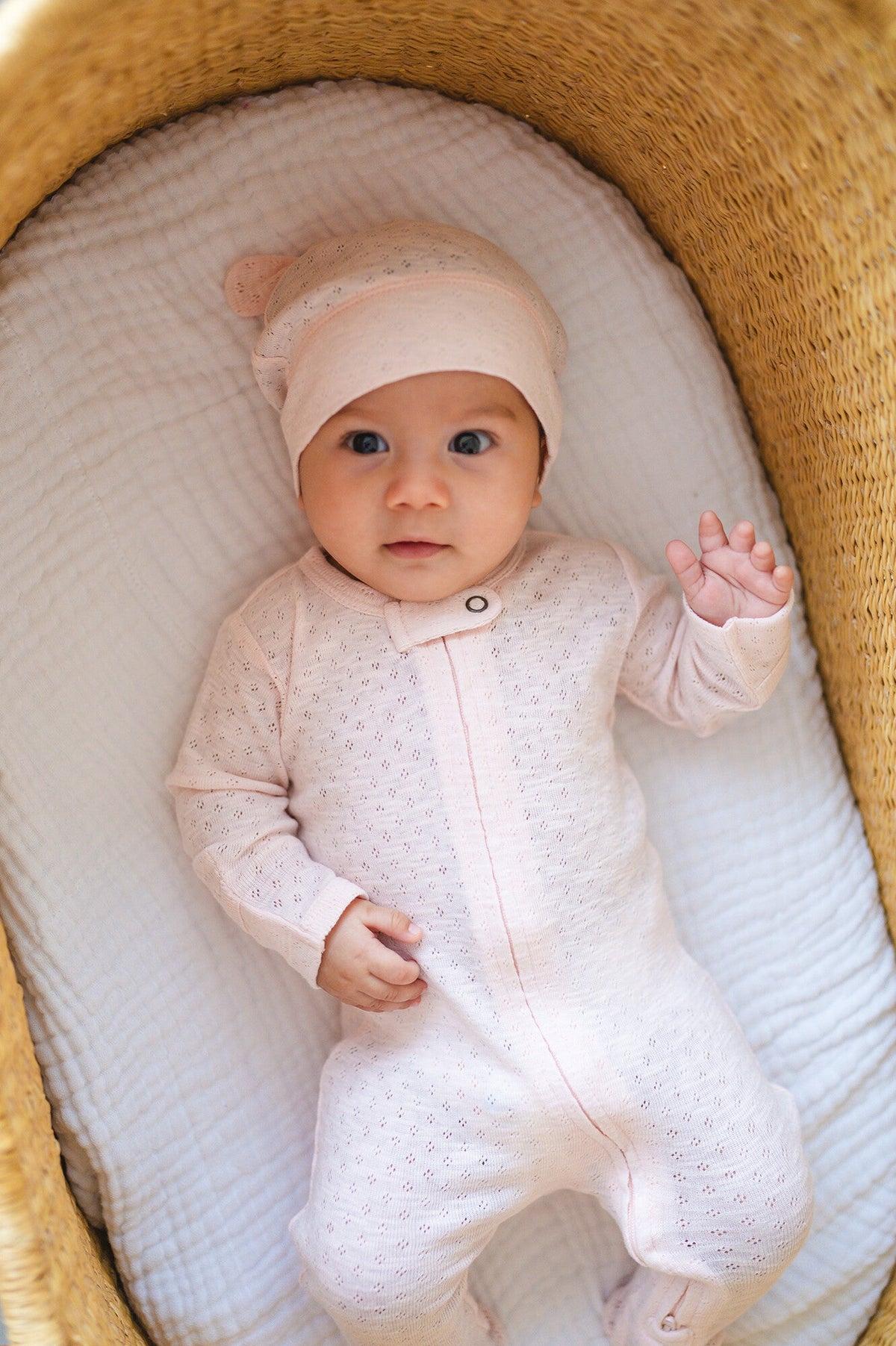 Baby in a white onesie and pink hat lying in a wooden crib.