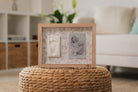 Wooden photo frame with baby milestone cards on a woven ottoman in a living room.
