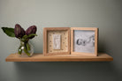 Wooden shelf with a vase of purple flowers and two wooden frames on a plain wall.