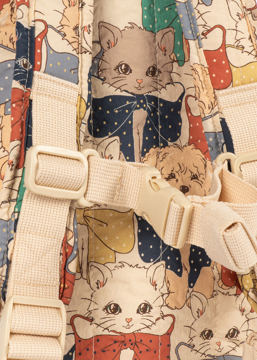 Close-up of a backpack featuring a whimsical print of cats and dogs in colorful clothing on a cream background, with beige webbing straps and plastic buckles prominently shown.