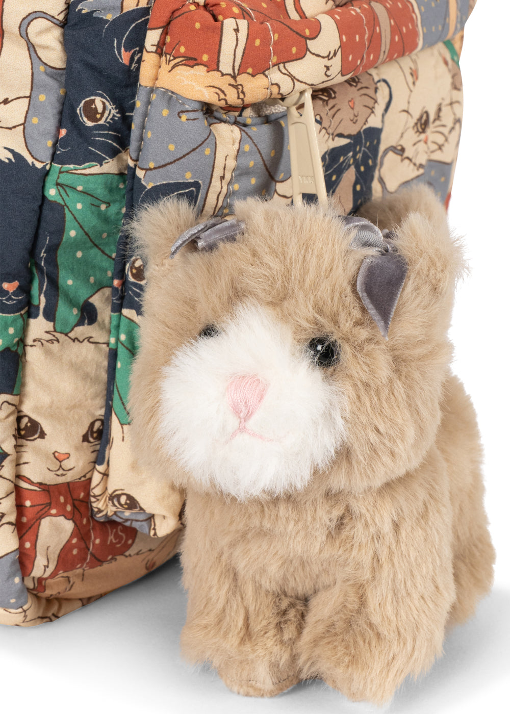 A plush toy kitten with fluffy tan fur, a white face, and a pink nose, posed in front of a patterned cat-print bag against a white background.