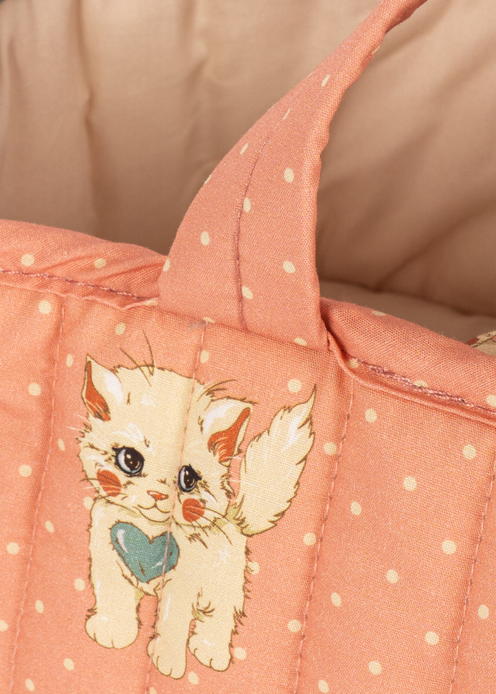 Close-up of a quilted peach-pink fabric item with white polka dots and an embroidered cartoon kitten holding a teal heart.