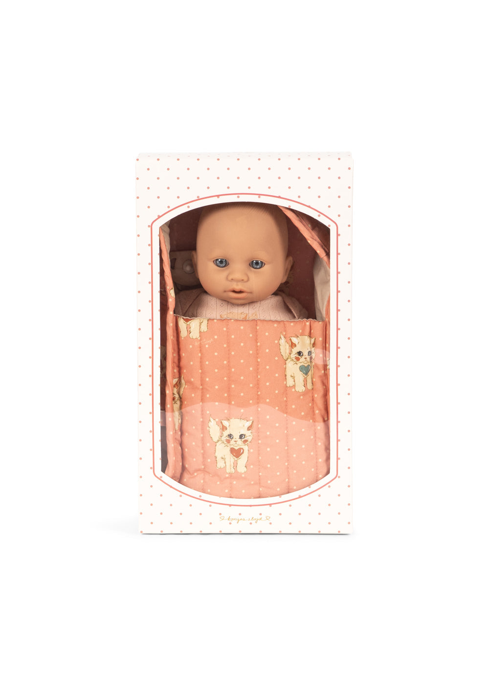 A doll with a bald head and blue eyes, wrapped in a pink blanket with white polka dots and small dog illustrations, presented in its original white box with a clear arched window.