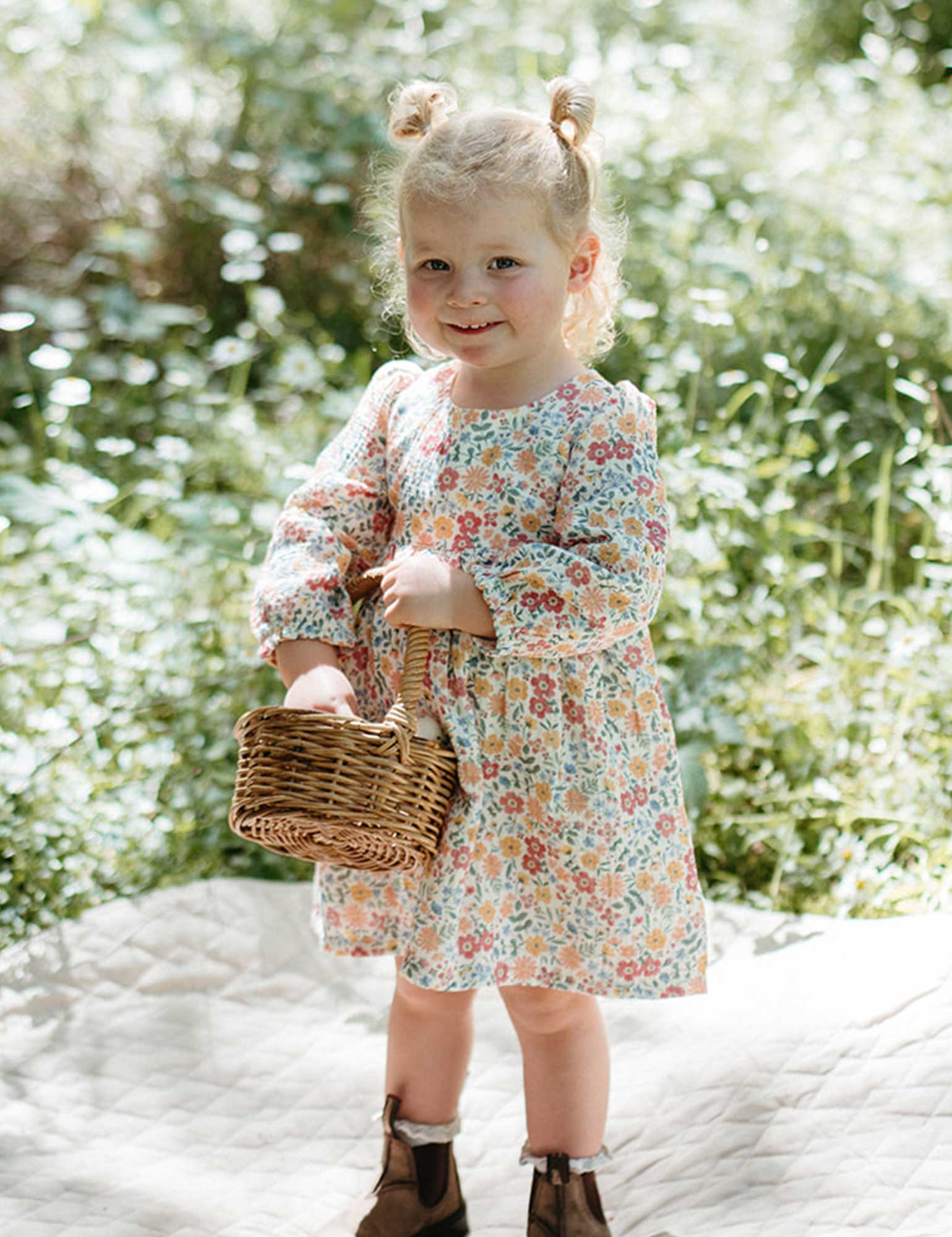 Young girl in a floral dress holding a basket outdoors with greenery in the background