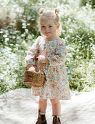 Young girl in a floral dress holding a basket outdoors with greenery in the background