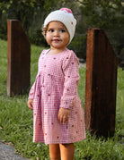 Child wearing a pink checkered dress and white hat with a pink bow outdoors.