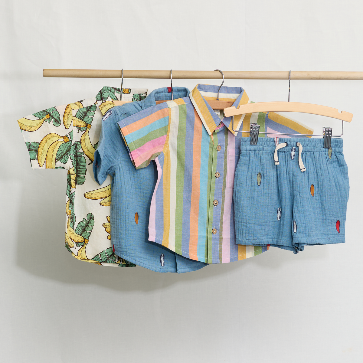 Children's clothing including a shirt with banana print, striped shirt, and blue shorts on a wooden rack against a white background.