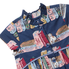 A close-up of a navy blue short-sleeve dress featuring a whimsical book-print pattern with illustrations of books, a cat, and text like 'Fairy Tales' and 'World Maps'. The dress has a ruffled mock neck, blue buttons, and a tiered skirt.