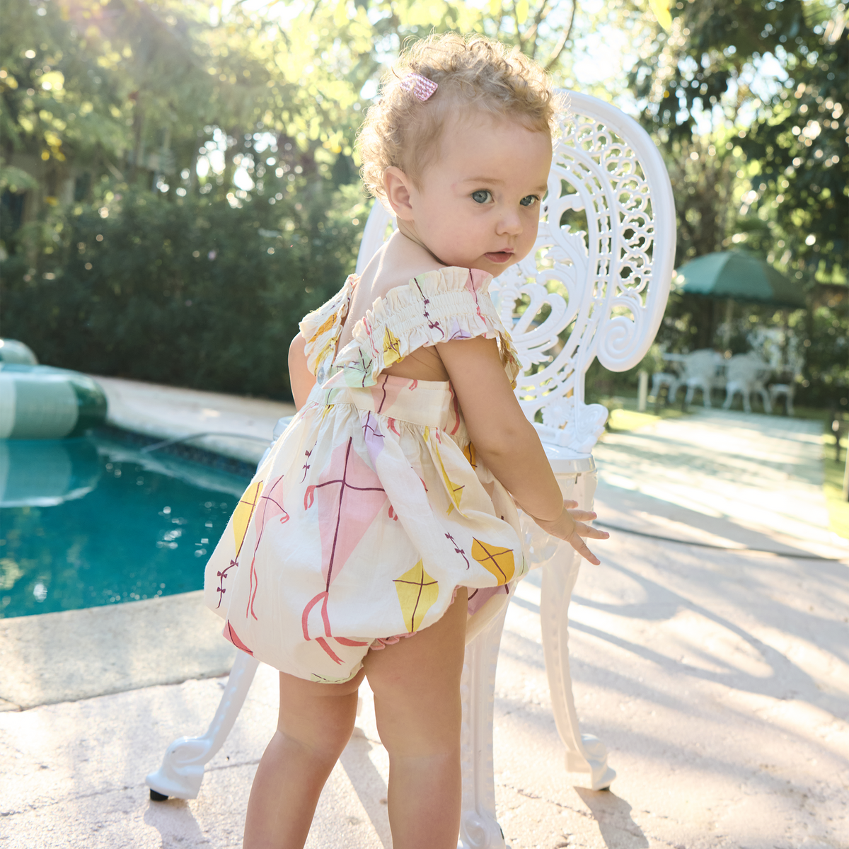 Child in a colorful dress standing by a poolside with trees in the background