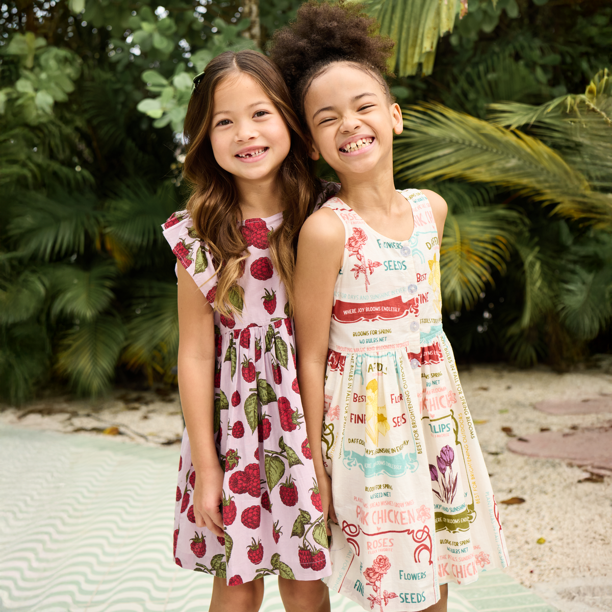 Two young girls wearing floral dresses standing outdoors with greenery in the background