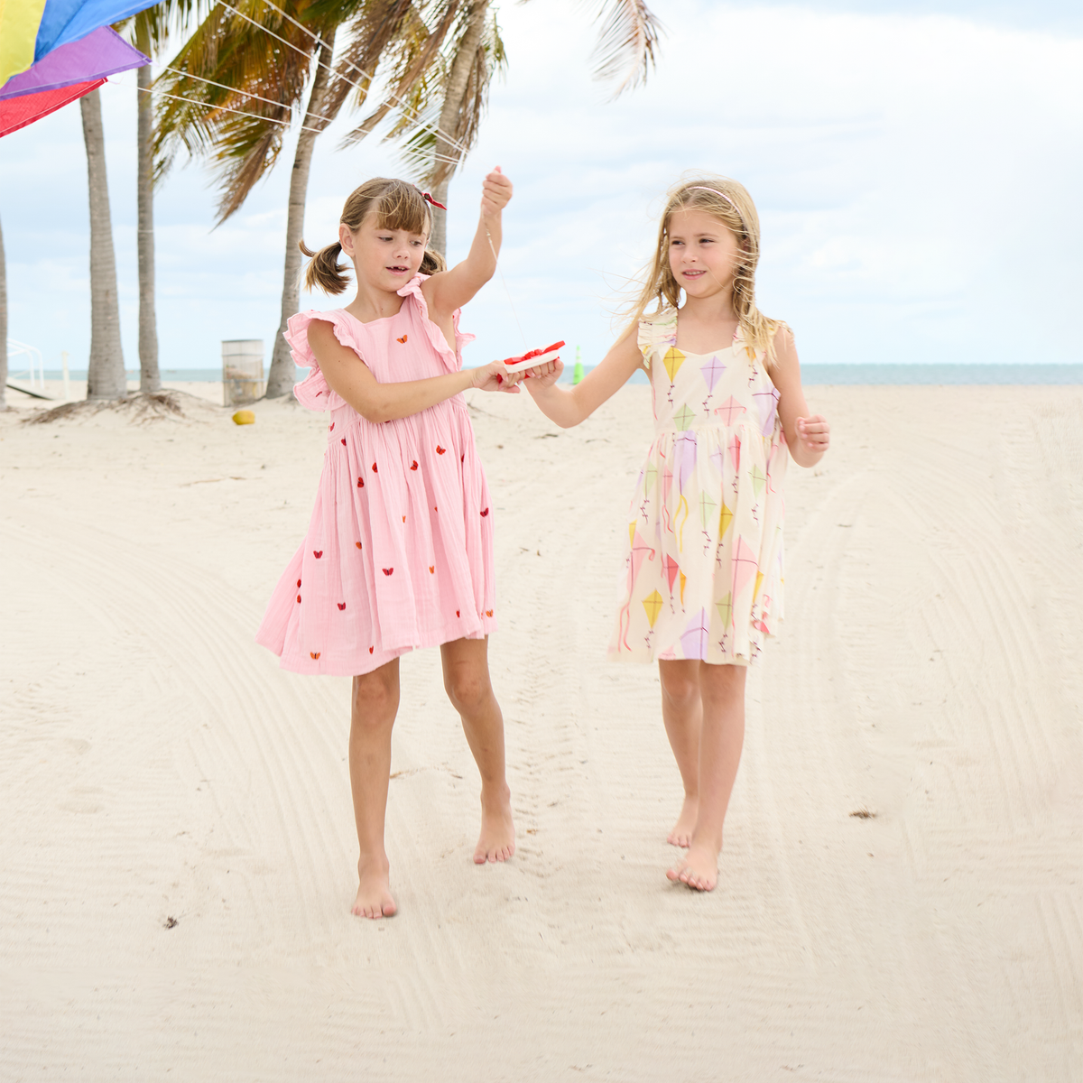 Two young girls in colorful dresses on a sandy beach with palm trees and a colorful kite in the background.