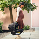 A pregnant woman smiling while carrying a black MIXX Next + Pipa Aire RX Travel System by Nuna infant car seat outdoors near a building and a tree.