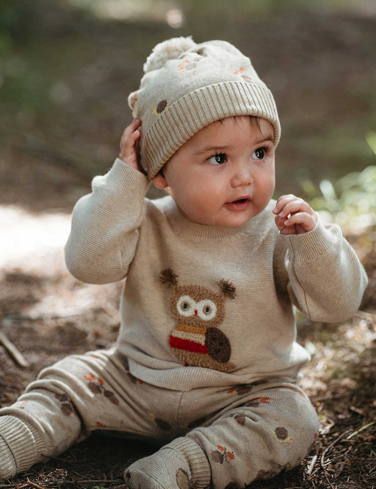 Baby wearing a beige knit outfit with an owl design, sitting on the ground outdoors.