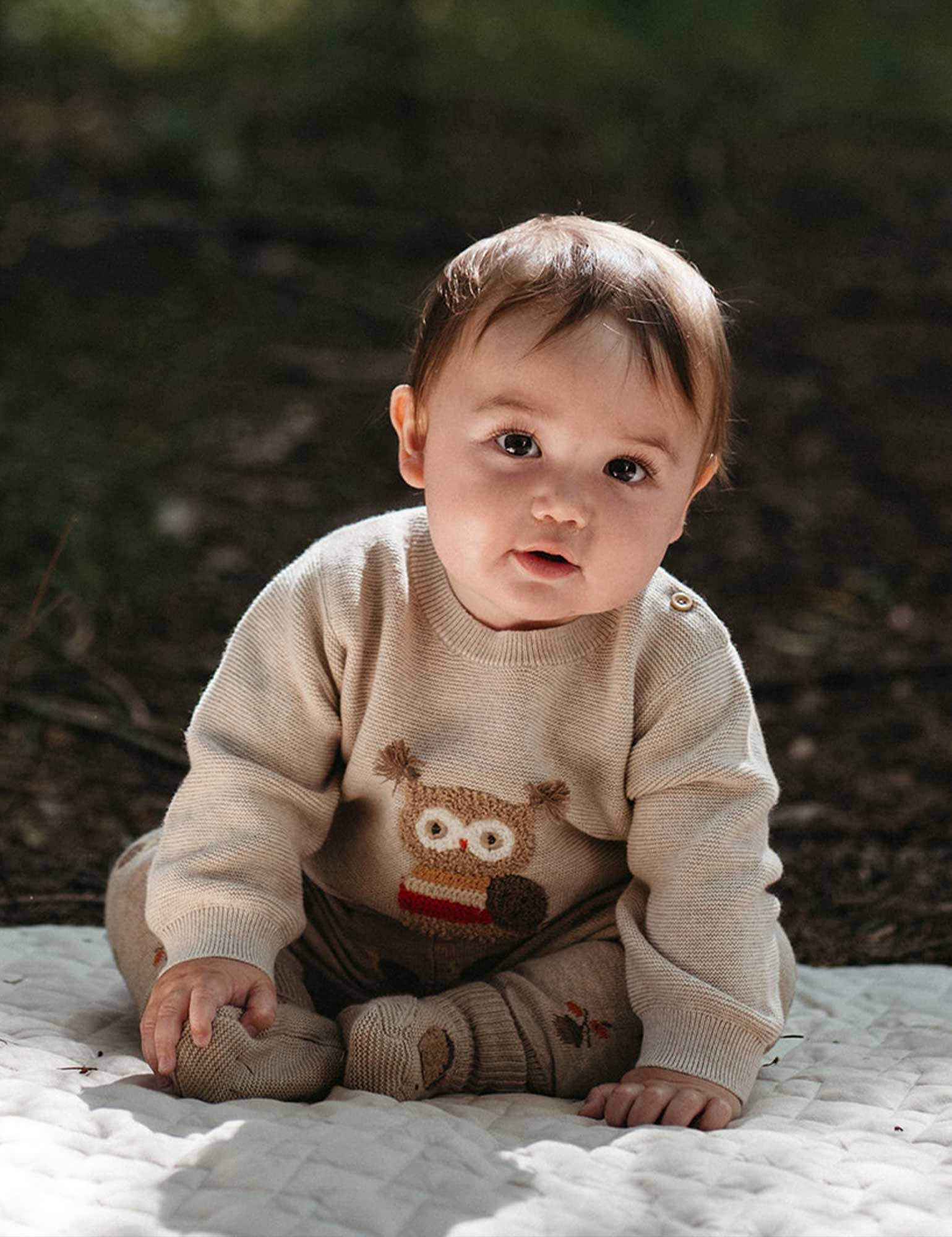 Baby wearing a beige sweater with an owl design, sitting on a blanket outdoors.
