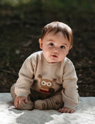 Baby wearing a beige sweater with an owl design, sitting on a blanket outdoors.
