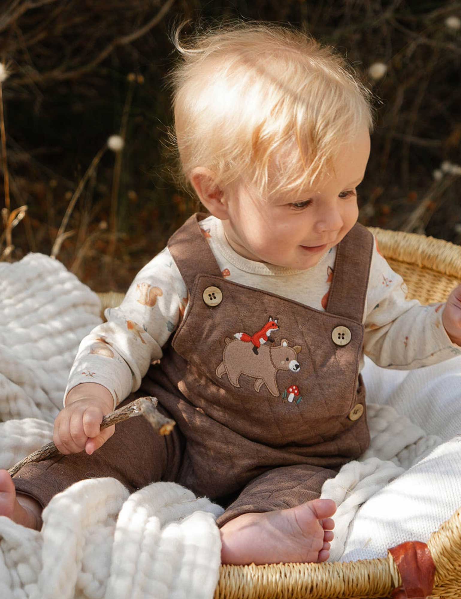 Baby in brown overalls sitting in a basket with a blurred natural background