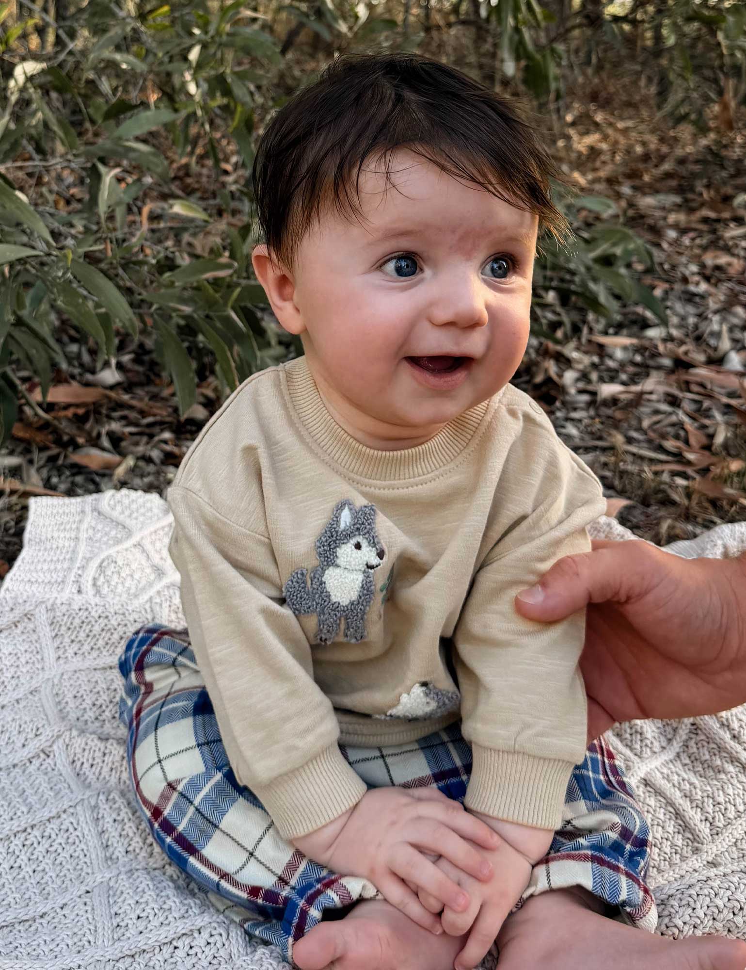 Baby wearing a beige sweater with a fox design, sitting outdoors on a blanket.