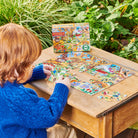A child with red hair wearing a blue sweater sitting at a wooden table outdoors, working on the 20 Piece Puzzle - Fairy Cottage by Eeboo with the puzzle box displayed in the background.