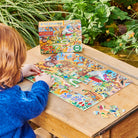 A child with red hair wearing a blue sweater sitting at a wooden table outdoors, working on the 20 Piece Puzzle - Fairy Cottage by Eeboo with the puzzle box displayed in the background.