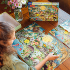 A young person sitting at a wooden table working on the 100 Piece Puzzle - Love of Nocturnal Life by eeBoo, with the puzzle box placed in the background next to a vase of flowers.