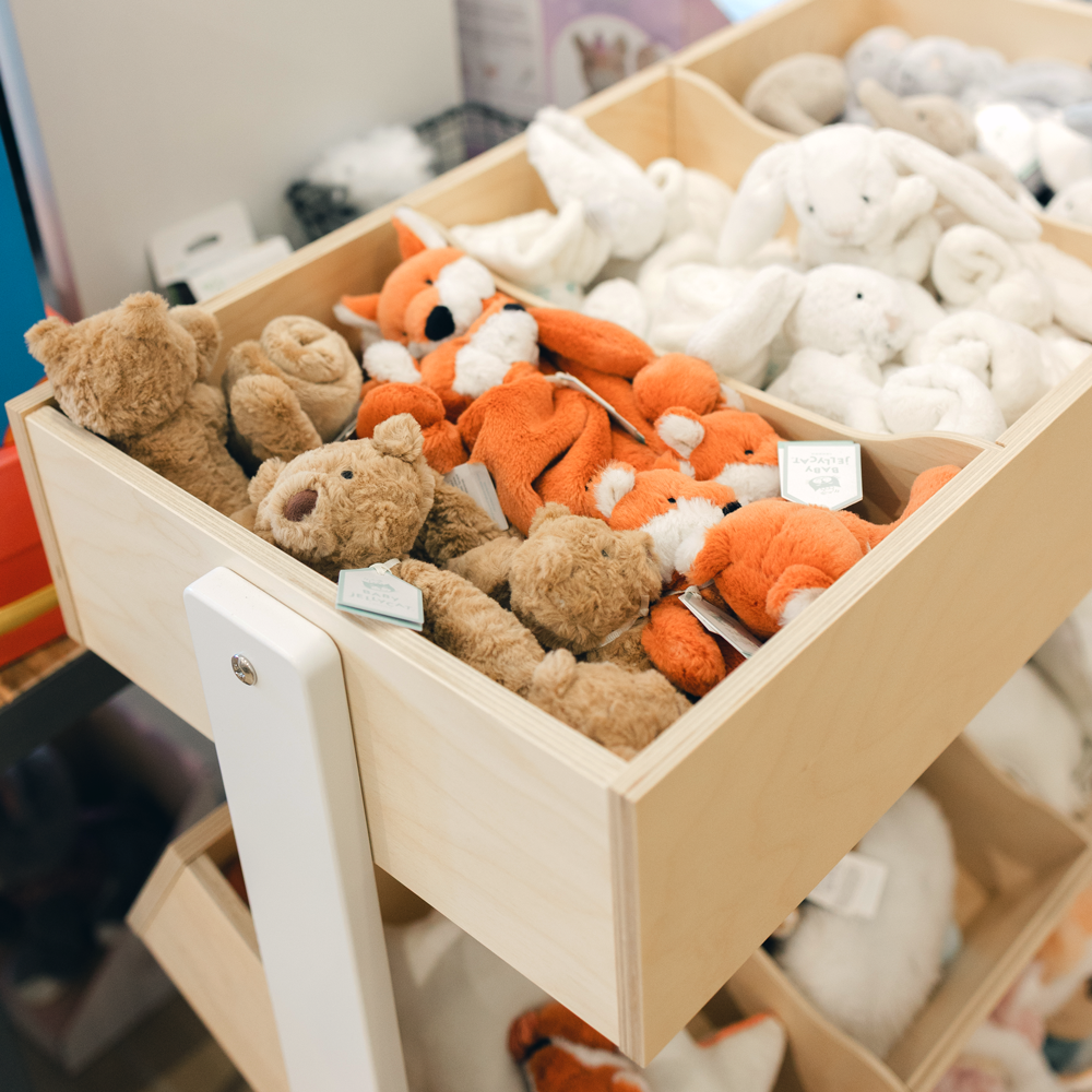 A wooden display drawer filled with plush stuffed toys, including brown teddy bears, orange foxes, and white bunny and dog figures, photographed in a store or playroom setting.