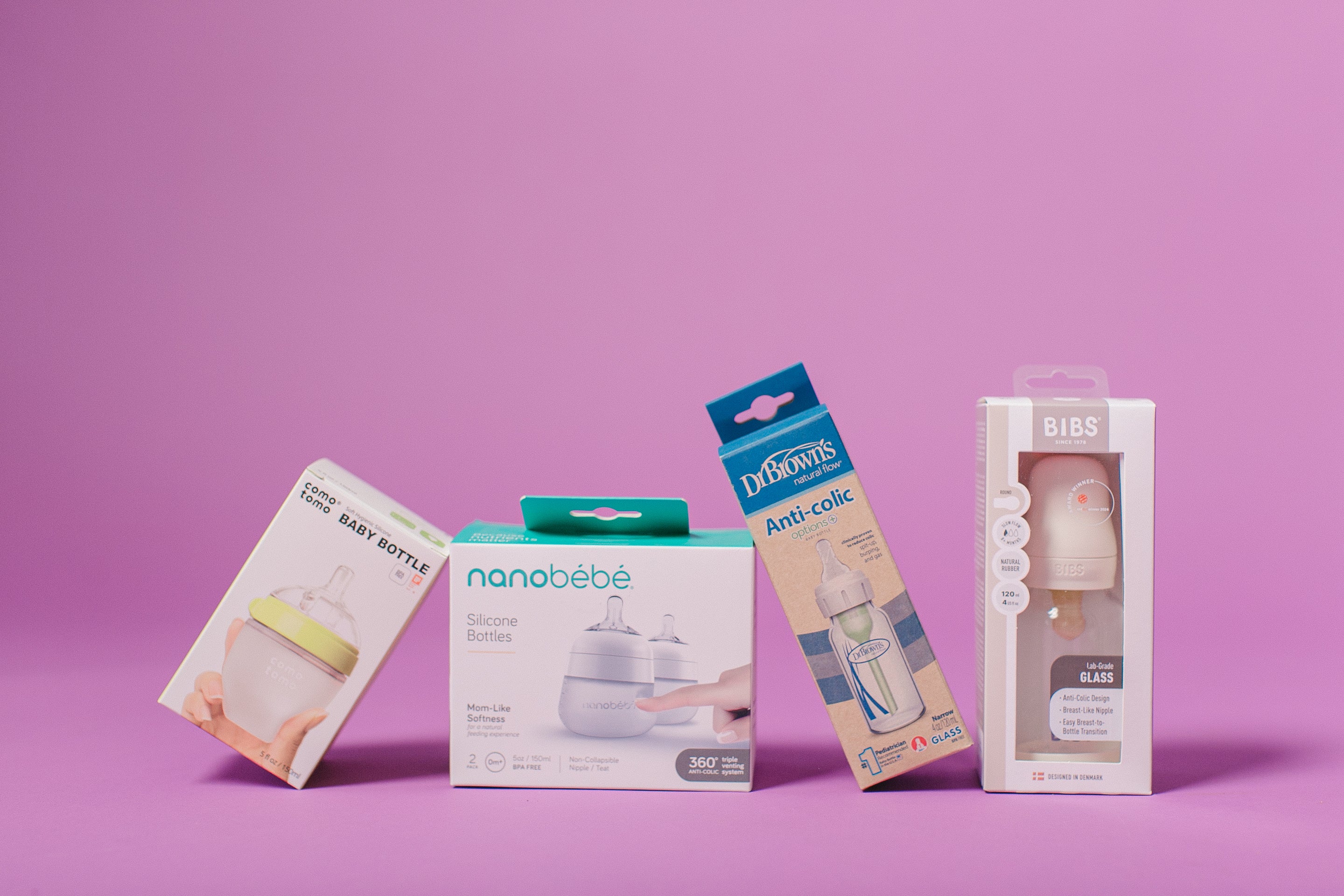 Four baby bottle packaging boxes arranged on a solid pink-purple studio background. The boxes include Little Baby Bottle, nanobébé Silicone Bottles, Dr.