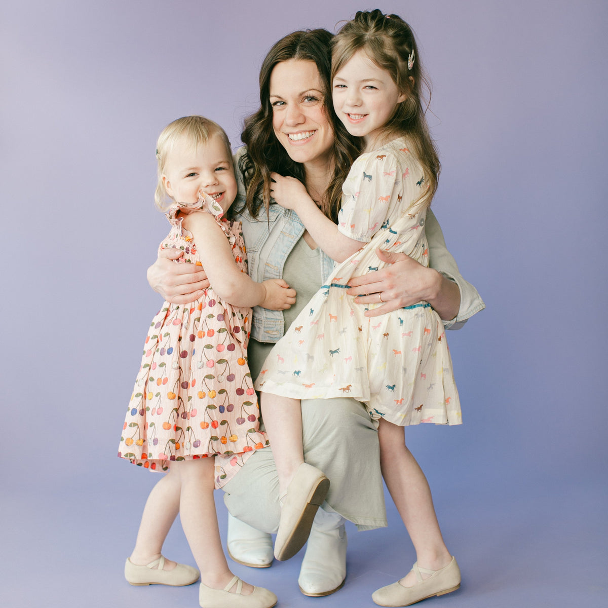 A family portrait of a mother and her two daughters, all smiling and posing together against a solid lavender studio backdrop. The girls are wearing matching patterned dresses, and the mother is wearing a light gray top and pants.