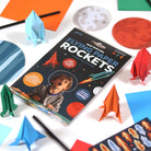 Book titled 'Flying Paper Rockets' surrounded by colorful paper rockets and craft materials on a white background