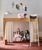 Two children sitting inside a wooden loft bed with white curtains, surrounded by colorful toys and books.