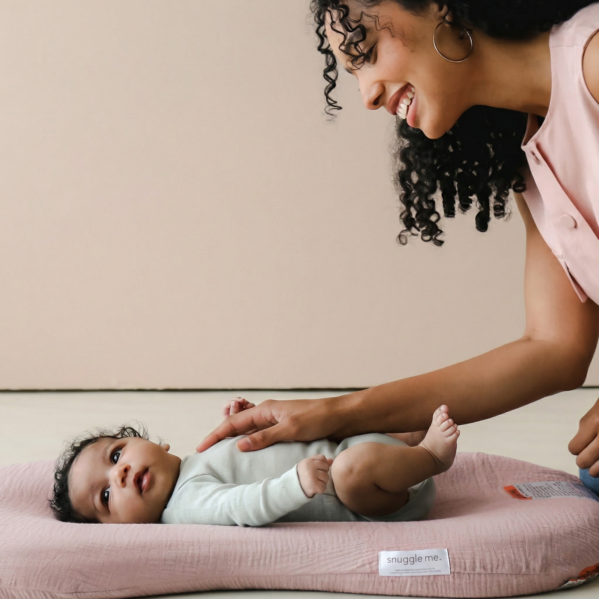 Woman interacting with a baby on a pink cushion against a beige wall.