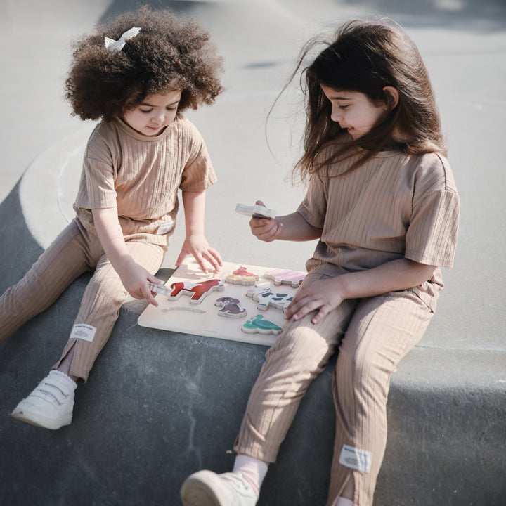 Two young girls wearing matching beige loungewear sets sitting outdoors on concrete steps, with one girl holding up a piece from a wooden farm animal puzzle while the other looks on.