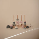 A wooden multi-shape stacker toy by Mushie & Co featuring four vertical pegs on a wooden base, with additional geometric wooden pieces scattered around the base in soft tones of cream, pink, sage green, and charcoal gray.