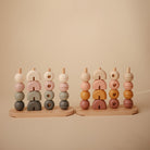 Two wooden multi-shape stacker toys by Mushie & Co standing side by side, featuring vertical pegs on wooden bases holding stacked geometric wooden pieces in earthy tones including cream, pink, sage green, and brown.