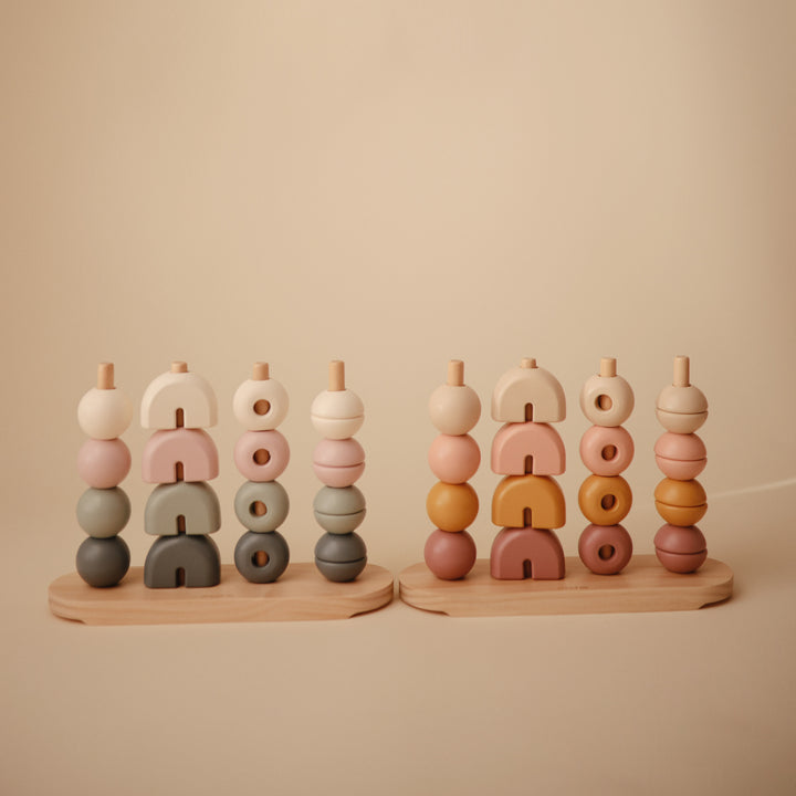 Two wooden multi-shape stacker toys by Mushie & Co standing side by side, featuring vertical pegs on wooden bases holding stacked geometric wooden pieces in earthy tones including cream, pink, sage green, and brown.