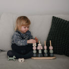 A toddler sitting on a bed playing with a wooden multi-shape stacker toy by Mushie & Co.
